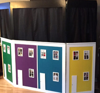 A four-panel puppet theatre that mimics a row of colourful townhomes. From left to right, the home panels are painted, green, violet, sky blue, and yellow.