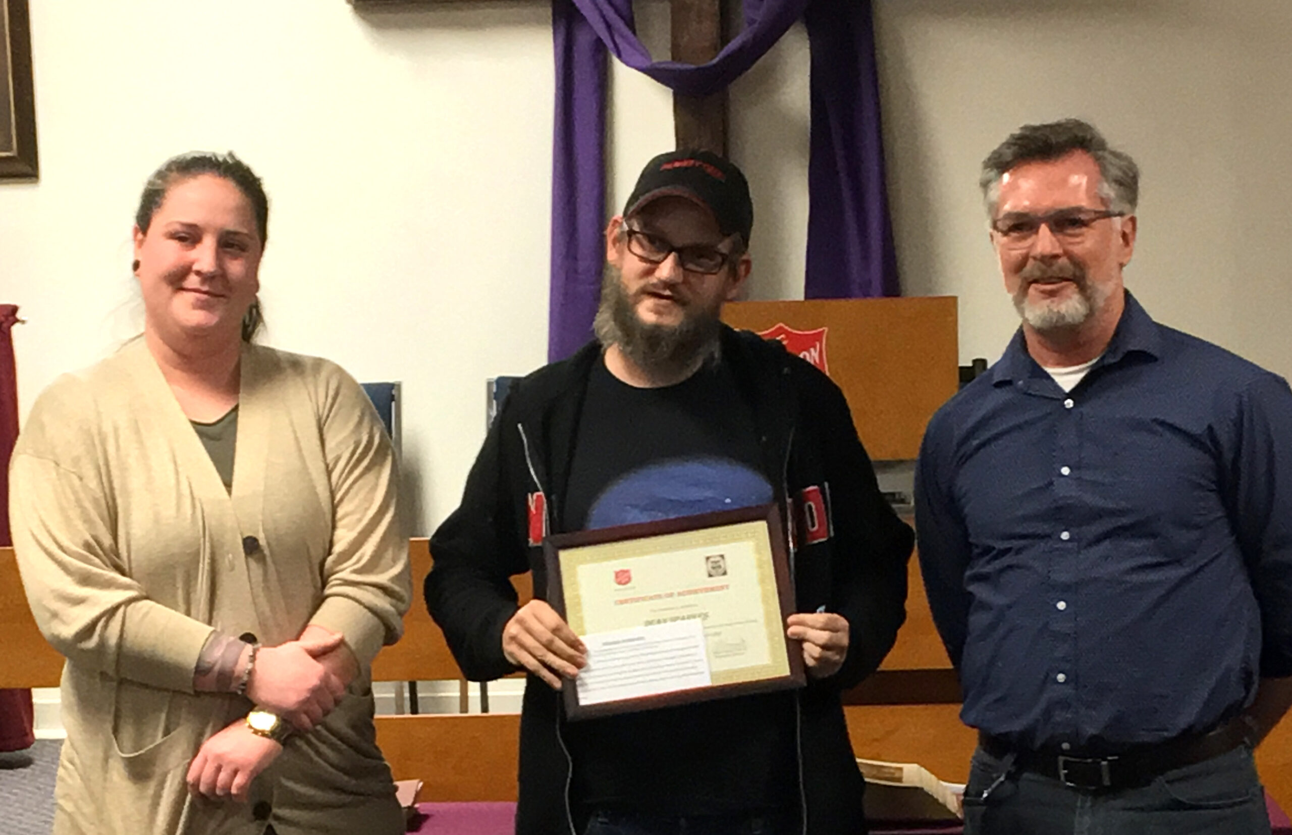 From left to right, a woman and two men stand at the front of the chapel and the man in the centre receives a certificate of completion for The Salvation Army Anchorage Recovery Program.