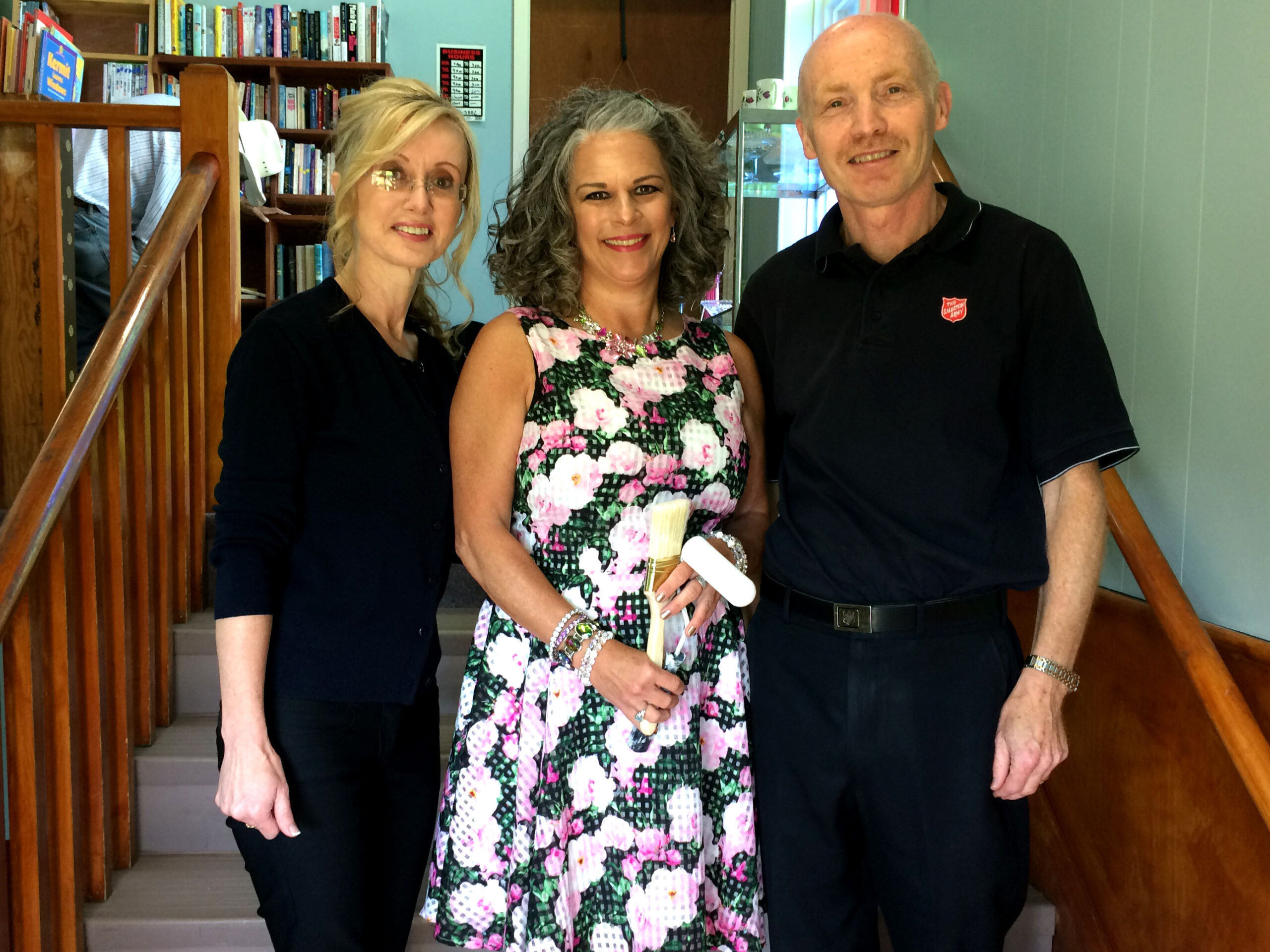 Two women and a man stand together for a photo. The women in the middle is wearing a flowered dress and is holding a paint brush. The woman on the left and the man on the right of her are both wearing black shirts and pants.