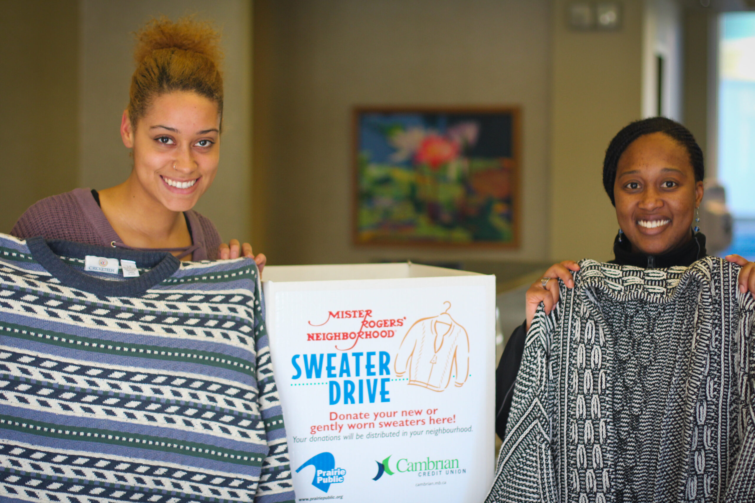 2 people holding up sweaters for Mister Rogers Sweater drive