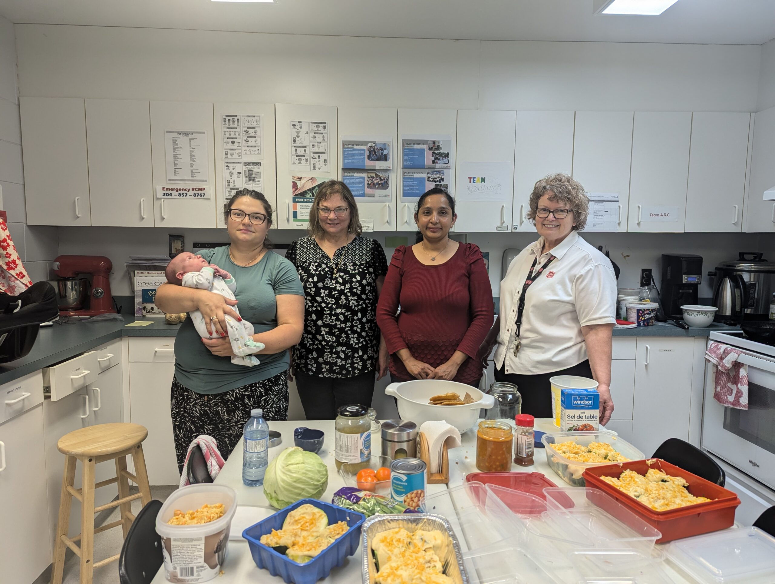 four women standing in a kitchen in front of meals that they have cooked.