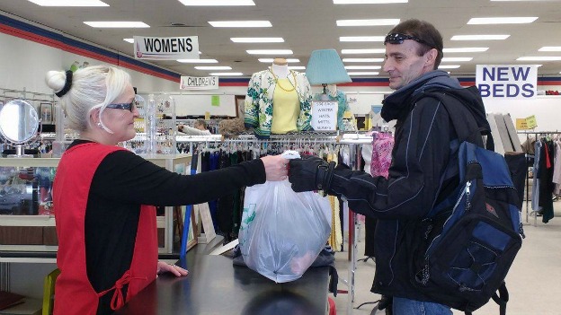 Thrift store worker stands behind cash register and hands bag of clothes to male customoer