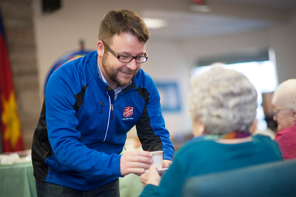 A Salvation Army employee handing a cup of tea to an elderly woman inside of a retirement home.