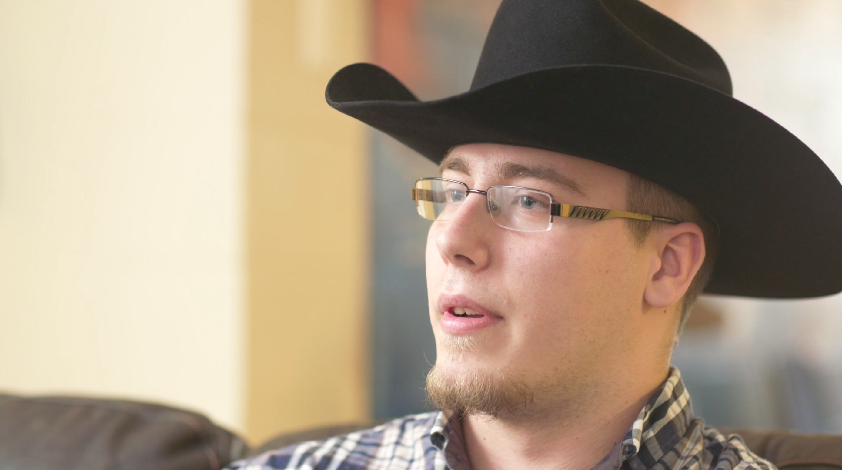 A closeup of a young man talking to a camera with a cowboy hat on and wearing glassed and a plaid shirt,