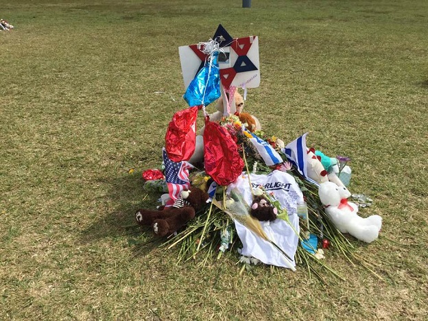 Photo of a memorial site with roses, an American flag, stuffed animals, and other flowers