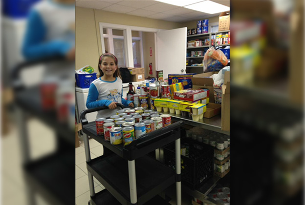 A young girl smiling at the camera, in front of her are many canned food items