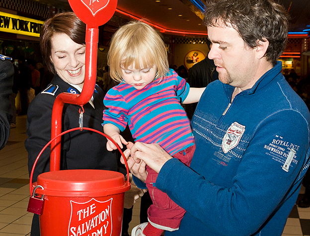 Salvation Army worker holds red kettle as father encourages child to insert coins