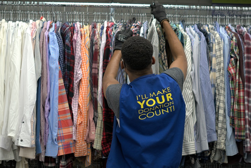 salvation army thrift store male worker hangs mens shirts while wearing a bib that says "I'll make your donation count"
