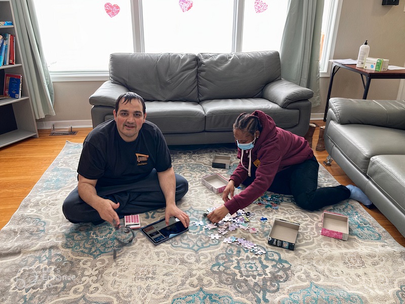 Sam smiles. Sits on floor with caregiver to piece together puzzle.