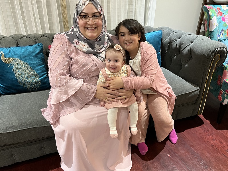 Elif sits on a couch holding her youngest daughter with her oldest daughter at their side