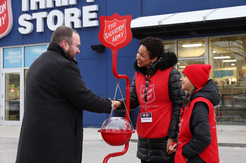 A man in a black coat and adult Salvation Army Christmas kettle volunteer, smiles and shake hands, a youth volunteer smiling to the side, Salvation Army thrift store in background.
