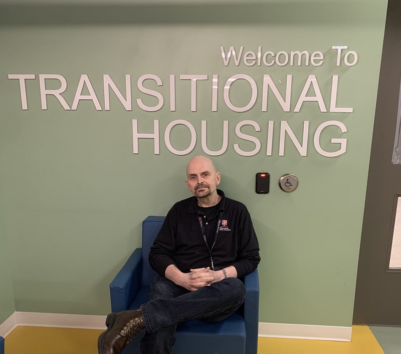 Craig sits on a chair in front of sign "Welcome to Transitional Housing."