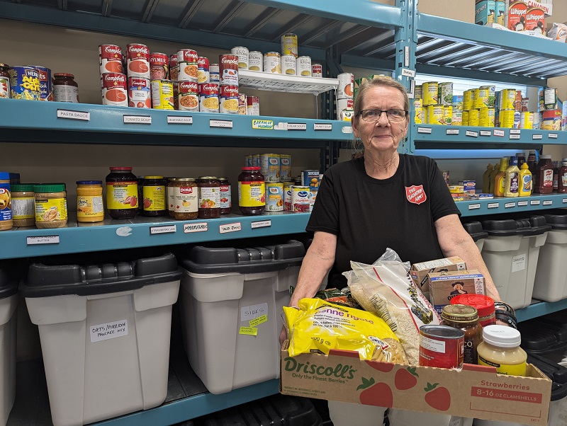 Salvation Army worker holds box of food while standing in front of food shelves