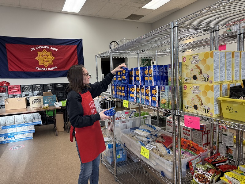 Salvation Army worker stands in front of shelves stocked with food. Not all shelves are filled.