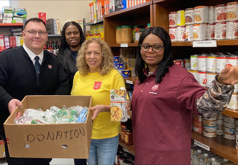Salvation Army staff stand in front of food bank shelves with box full of food for distribution