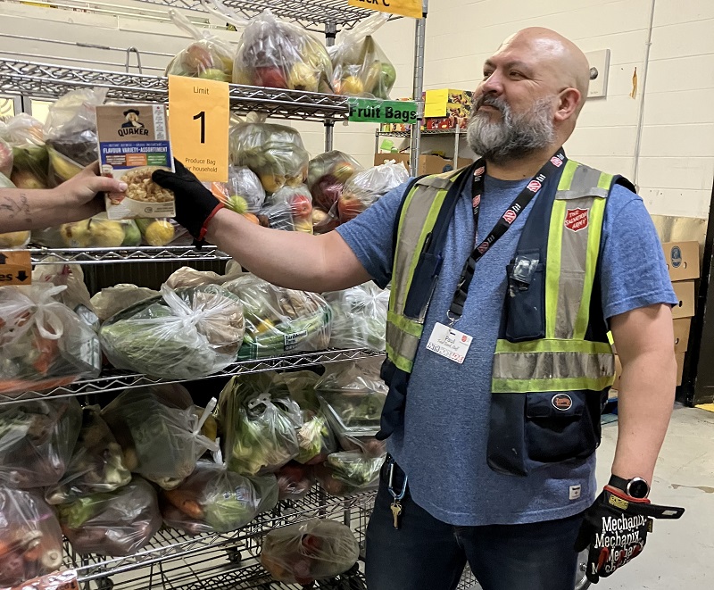 Salvation Army food bank worker stands in front of fresh food items and hands client box of cereal